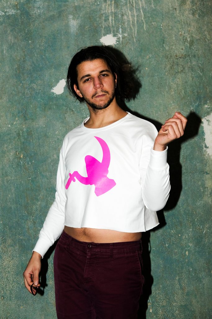 Ein Model mit langen Haaren und Bart trägt ein Crop-Top mit einem sehr großen Auftdruck mit Buttplug-und-Sichel-Motiv auf der Brust.